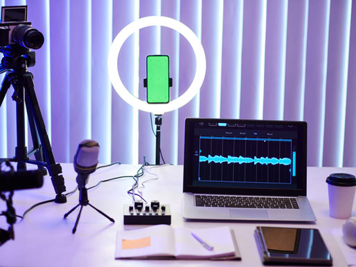 A content creation setup with a camera on a tripod, a ring light holding a smartphone with a green screen, and a laptop displaying audio editing software on a desk. A microphone, audio mixer, notebook, tablet, headphones, and a takeaway coffee cup are arranged neatly, with soft neon lighting in the background.