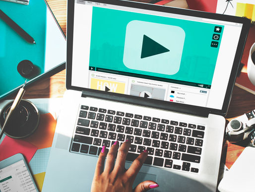 Hand with pink nails types on a laptop with a large play button on screen. Desk is cluttered with stationery, a phone, and a coffee cup.