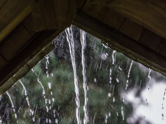 Rain pours through a wooden roof opening, streams and droplets falling against a blurred green-and-white background.