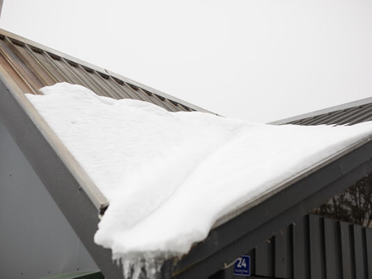 Snow piled thickly on a gray metal roof under an overcast sky, with a small blue 24 sign below.