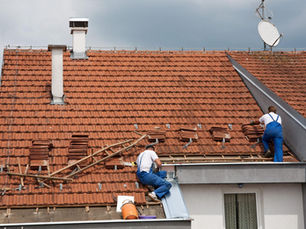 Two workers in blue overalls repair a red tile roof. One measures, while the other arranges tiles. Overcast sky in the background.