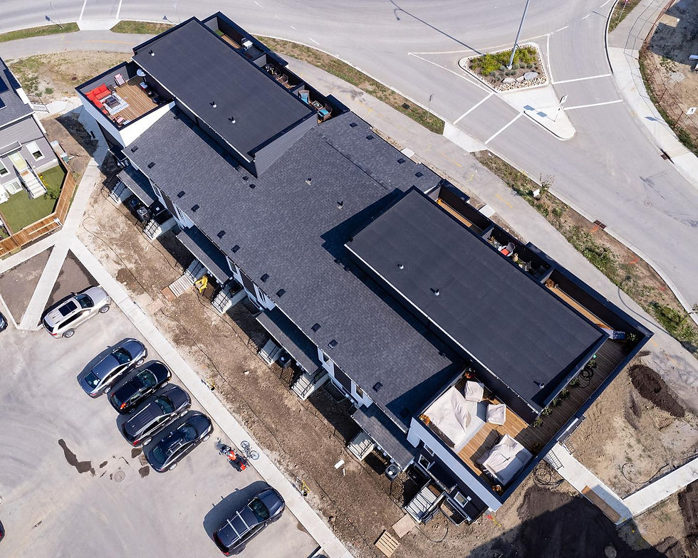 Aerial view of a modern black-roofed building with rooftop patios. Six parked cars and a bike are visible in the adjacent lot. Roads surround.