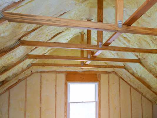 Attic interior with spray foam insulation and exposed wooden rafters above a bright window, showing unfinished construction