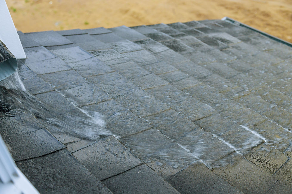 Water rushing off a gray shingled roof onto the ground, with a gutter downspout at left and sandy background