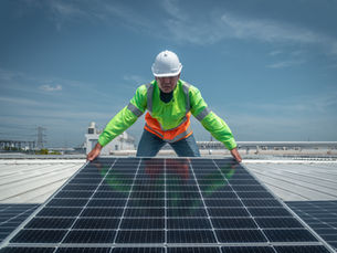 A worker in a hard hat installs a solar panel on a rooftop, with blue sky and industrial buildings behind.