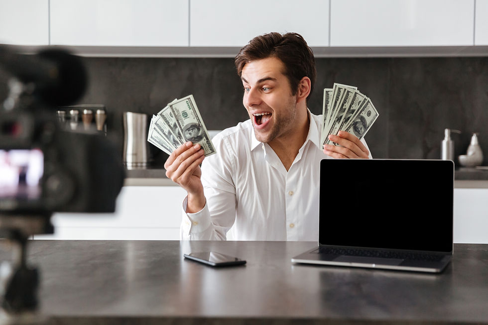 Excited man in a kitchen holds fanned $100 bills while filming with a camera; laptop and phone on counter, no screen text visible