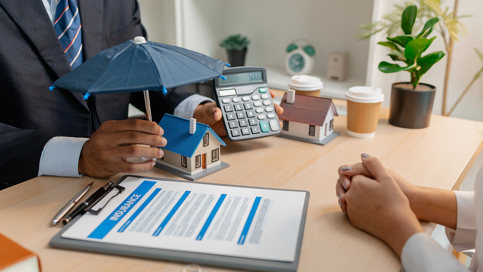 Insurance agent with calculator and model houses under umbrella, meeting client at desk with policy paperwork in bright office.