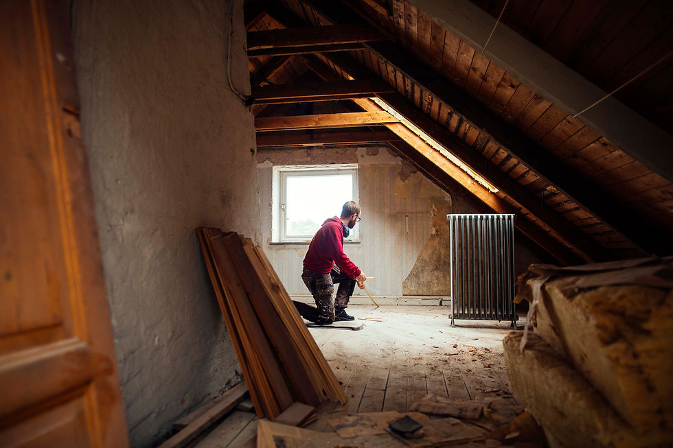 Man in red hoodie kneels in a dusty attic renovation, holding a tool near a window under exposed wooden beams.