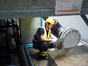 Worker in yellow hard hat and reflective vest crouches with a laptop beside large industrial pipes.