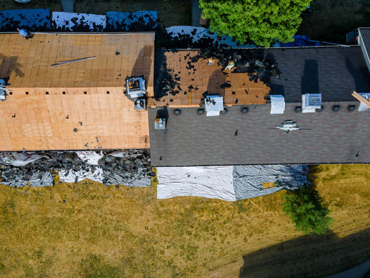 Aerial view of roofers stripping and replacing shingles on a house, with tarps, tools, and a leafy tree beside the roof.