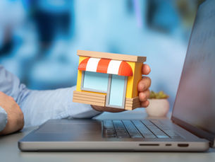 Hand holding a small storefront model beside an open laptop, suggesting online retail or e-commerce work.