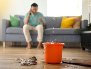 Orange bucket catches a ceiling leak on a wet living room floor while a man sits on a sofa talking on the phone.