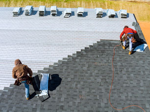 Two workers install gray roof shingles on a house; stacks of shingles are visible. One uses a nail gun. Clear, sunny day.