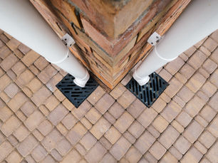 Top-down view of a brick pillar corner with two white downspouts draining into black grates on tan paver stones.