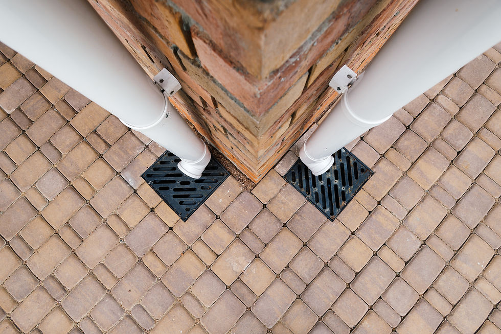 Top-down view of a brick pillar corner with two white downspouts draining into black grates on tan paver stones.