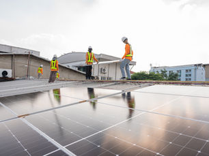 Workers in hard hats and safety vests install solar panels on a rooftop under a bright sky.