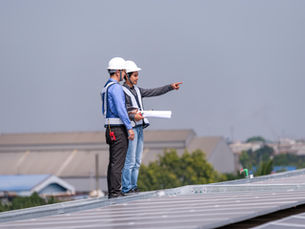 Two engineers in hard hats on a rooftop, one pointing while holding blueprints, with solar panels in foreground and cloudy sky.