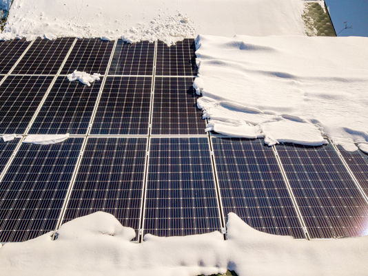 Snow-covered solar panels on a rooftop, with dark panels exposed in patches under bright winter sunlight.