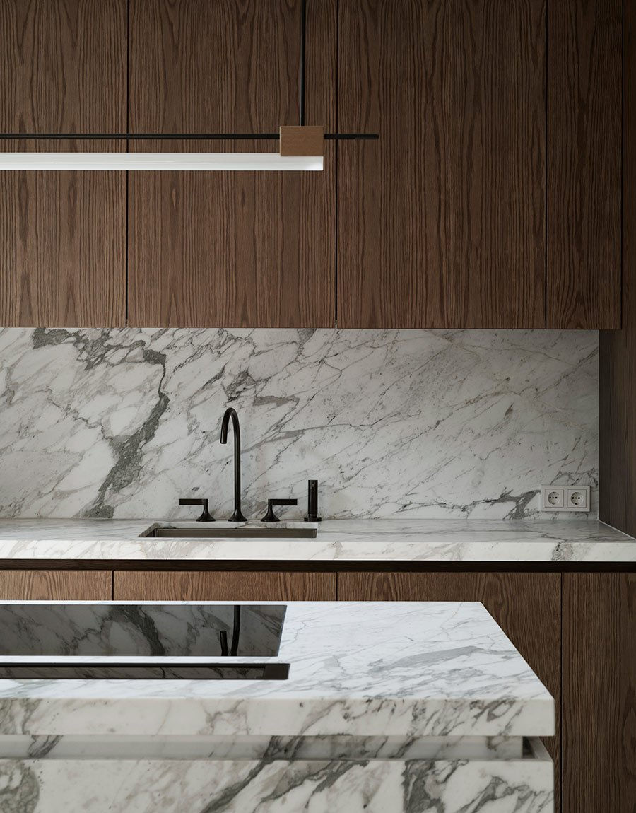 A detailed view of a modern kitchen's section, showcasing the harmonious blend of natural wood cabinetry with striking marble countertops and backsplash. The streamlined design is accentuated by a sleek, black faucet and minimalist shelf lighting, creating a sophisticated and serene cooking space that emphasizes clean lines and high-quality materials.