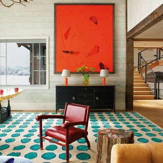 A vibrant interior featuring a large orange geometric artwork above a black console, red armchair, and a turquoise rug with circular patterns.