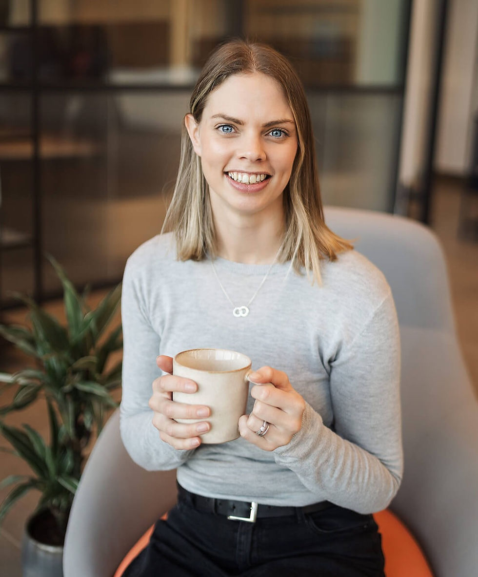 Charlotte, a white woman with blonde hair wears a grey jumper and is sitting in a chair with a cup in her hands smiling at the camera.