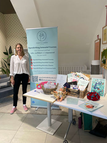 A woman stands indoors in a John Lewis nursery department next to a table, smiling and presenting a display for Positive Birth Leeds. Next to her is a branded banner that reads, "Positive Birth Leeds: Hypnobirthing Antenatal Courses – Get informed and feel calm, confident, and empowered for your birth!" Below it are listed services such as "Hypnobirthing Antenatal Courses," "Hypnobirthing Essentials Courses," "Hypnobirthing Refresher Classes," and "Hypnobirthing for a Caesarean Birth," with options for group, private, and online courses. A table in front displays educational materials, printed guides, tote bags, and birthing resources, arranged neatly.