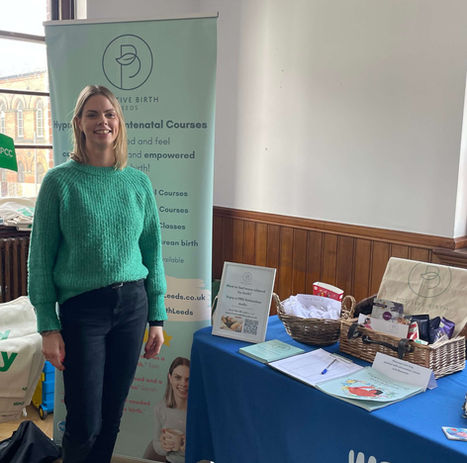 A woman stands indoors, smiling and presenting a display for Positive Birth Leeds. Behind her is a branded banner that reads, "Positive Birth Leeds: Hypnobirthing Antenatal Courses – Get informed and feel calm, confident, and empowered for your birth!" Below it are listed services such as "Hypnobirthing Antenatal Courses," "Hypnobirthing Essentials Courses," "Hypnobirthing Refresher Classes," and "Hypnobirthing for a Caesarean Birth," with options for group, private, and online courses. A table on her right displays a gift basket and educational materials, printed guides, tote bags, and birthing resources, arranged neatly.