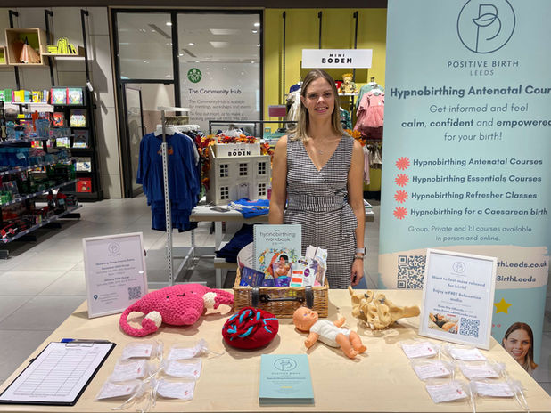 A woman stands indoors in a John Lewis clothing department behind a table, smiling and presenting a display for Positive Birth Leeds. Next to her is a branded banner that reads, "Positive Birth Leeds: Hypnobirthing Antenatal Courses – Get informed and feel calm, confident, and empowered for your birth!" Below it are listed services such as "Hypnobirthing Antenatal Courses," "Hypnobirthing Essentials Courses," "Hypnobirthing Refresher Classes," and "Hypnobirthing for a Caesarean Birth," with options for group, private, and online courses. A table in front displays educational materials, printed guides, tote bags, and birthing resources, arranged neatly.