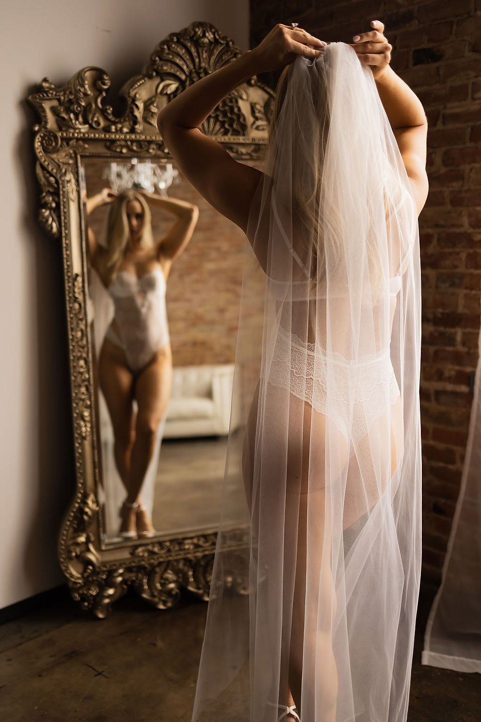 This is a bridal boudoir photograph taken by the boudoir photographer JoAnna Moore of a bride in white bridal lingerie looking at herself in the mirror. The bride is holding onto her wedding veil.