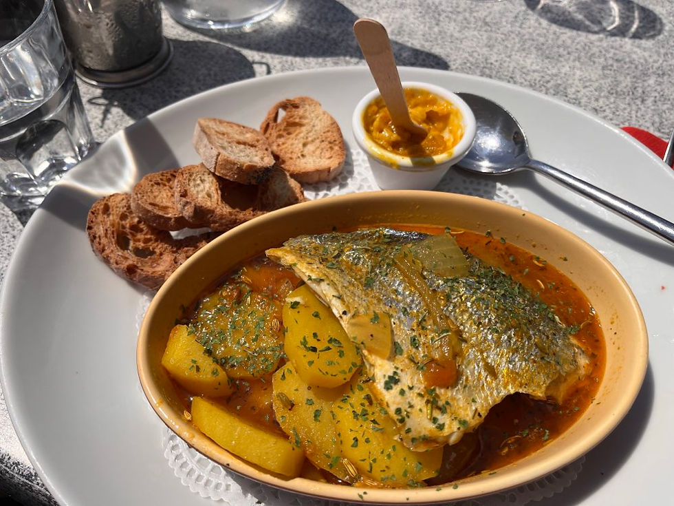 Gegrillter Fisch mit Kartoffeln in roter Sauce, dazu Brotscheiben und Dip auf einem weißen Teller im Sonnenlicht.