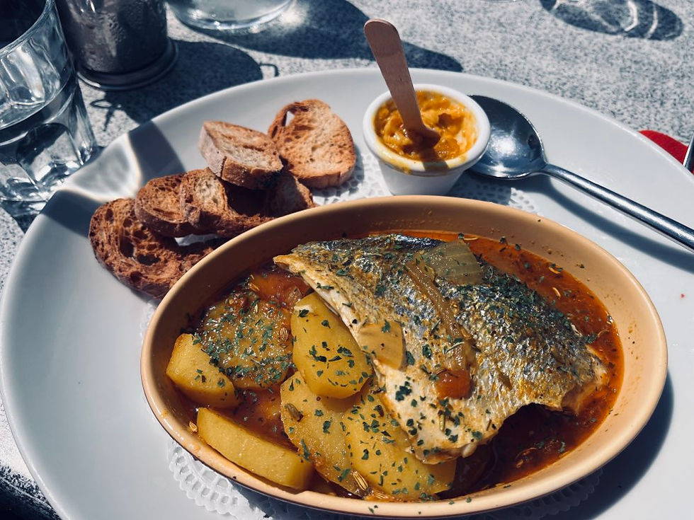 Fischteller mit Kartoffeln und Tomatensauce, garniert mit Kräutern. Im Hintergrund Brot, ein Glas und eine Schale mit gelbem Dip.