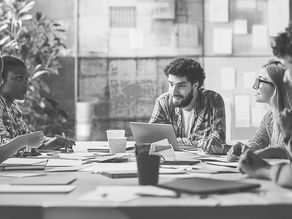 Five people in a meeting room discussing Brand Activations around a table, papers and laptops visible. Black and white setting with a collaborative mood.
