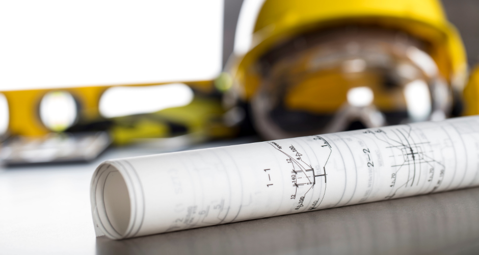 Rolled architectural blueprint on desk, with blurred yellow hard hat and level in background, suggesting construction planning.