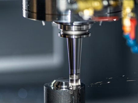 A CNC machine precisely mills a metal piece, sparking slight shavings. Background is industrial with yellow and blue components visible.