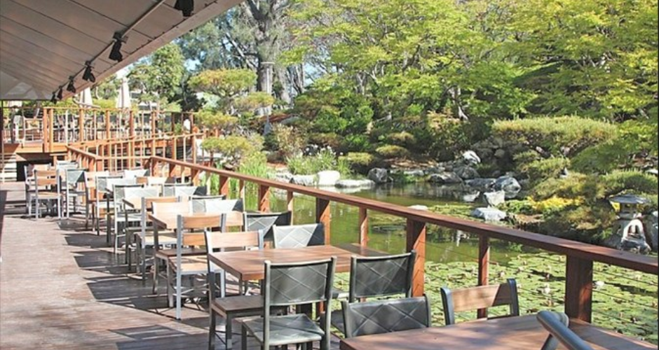 Outdoor cafe with empty tables and chairs on a wooden deck overlooking a lush, green Japanese garden with a pond reflecting the serene setting.