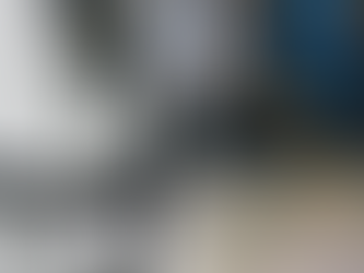 Close-up of a CNC machine head with a drill bit. Blue and orange coolant hoses are visible. Background features a metal surface.