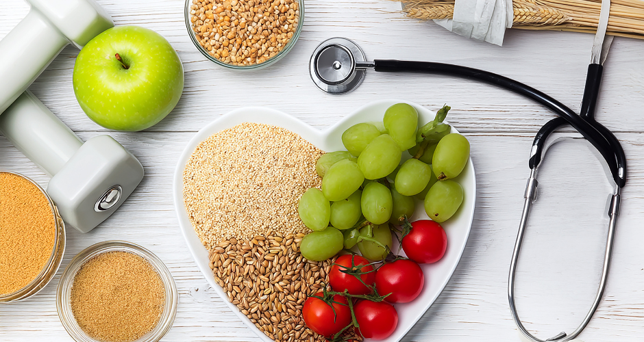 Heart-shaped bowl with grains, grapes, and tomatoes on a white surface. Nearby are a green apple, dumbbells, and a stethoscope. Health concept.