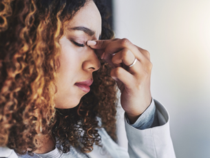 Woman with curly hair pinches her nose bridge, eyes closed, in a bright setting. She appears stressed or tired, wearing a light jacket.