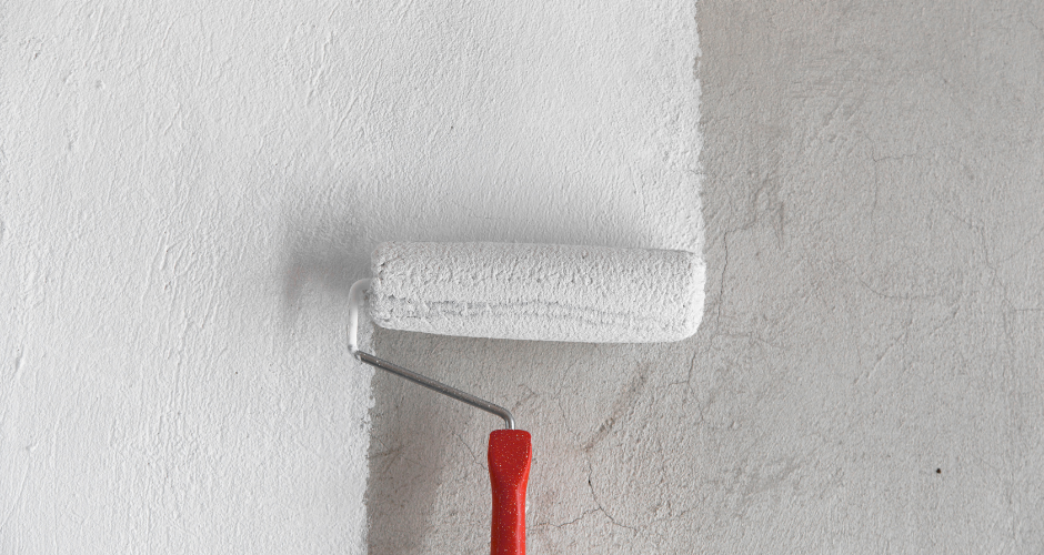 A paint roller with a red handle applies white paint on a gray wall, splitting the wall into painted and unpainted sections. Minimalist setting.