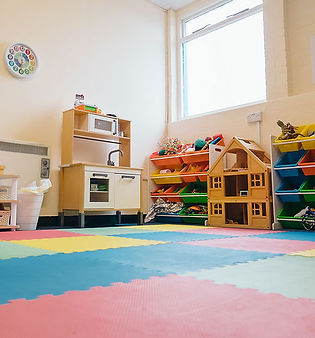 A spacious, sunlit room with multi-colored interlocking floor mats and various play-based therapy stations.