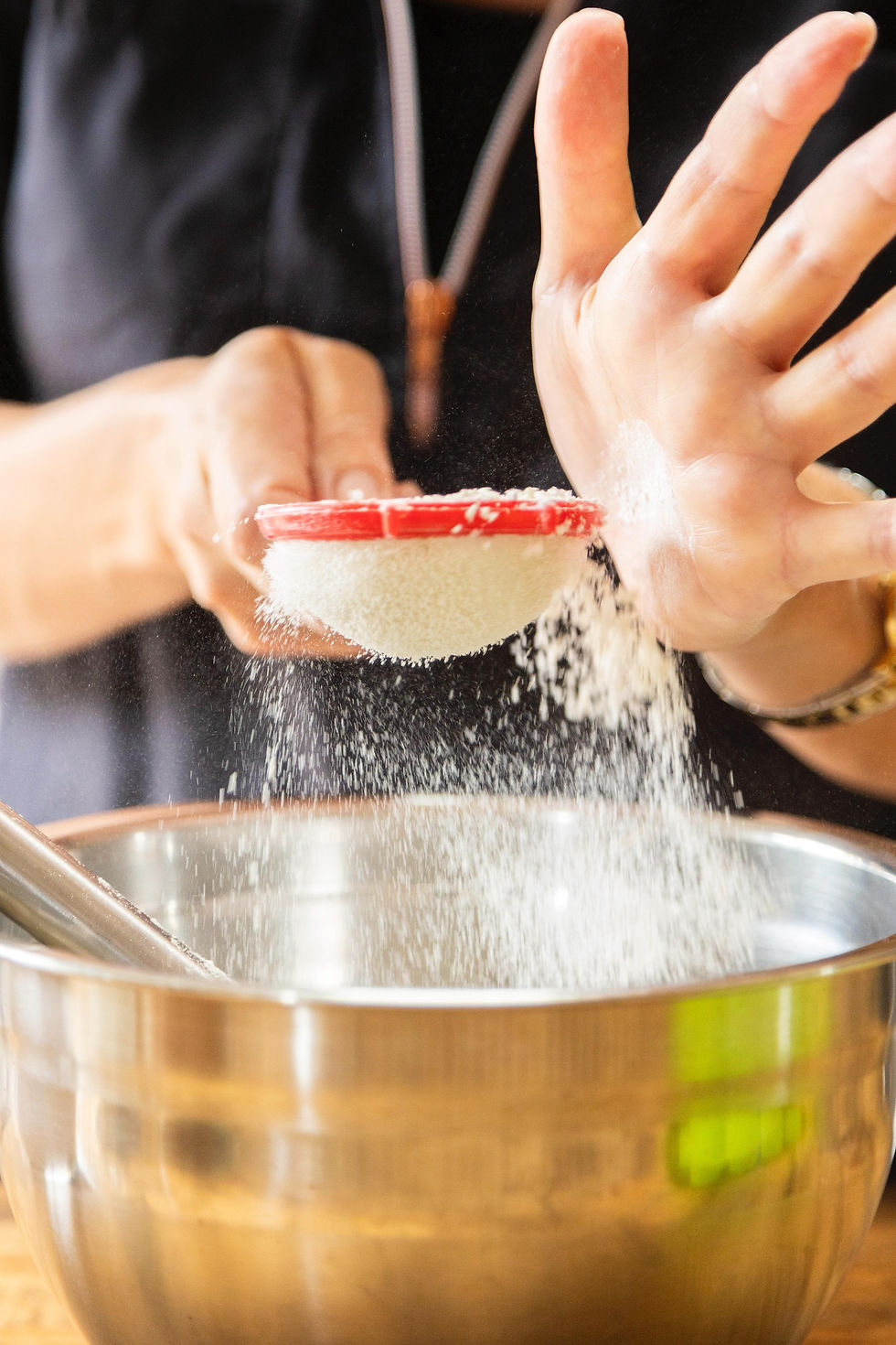 Mains de Cécile tamisant de la farine dans un saladier inox en préparation de cupcakes artisanaux