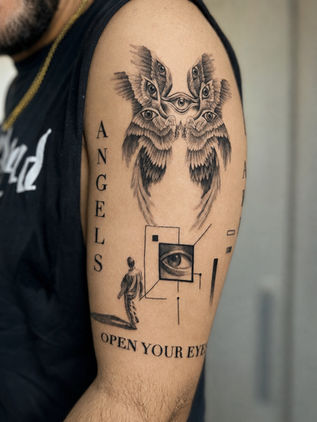 Black and grey surreal upper arm tattoo featuring angel wings, multiple realistic eyes, geometric linework, and typography, tattooed at White Sloth Tattoo Shop in Tamarindo, Costa Rica.