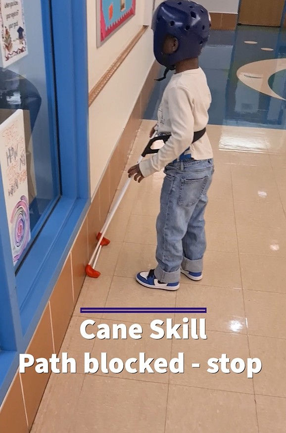 A child wearing a helmet wears a belt cane in a school hallway, facing a wall. Text reads "Cane Skill: Path blocked - stop."