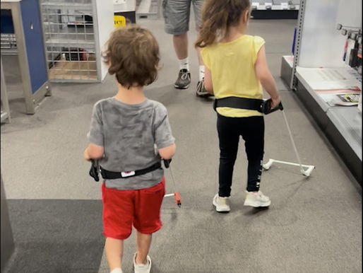 Two children walk in an electronics store wearing Belt Canes, one in red shorts and one in yellow, with an adult ahead.