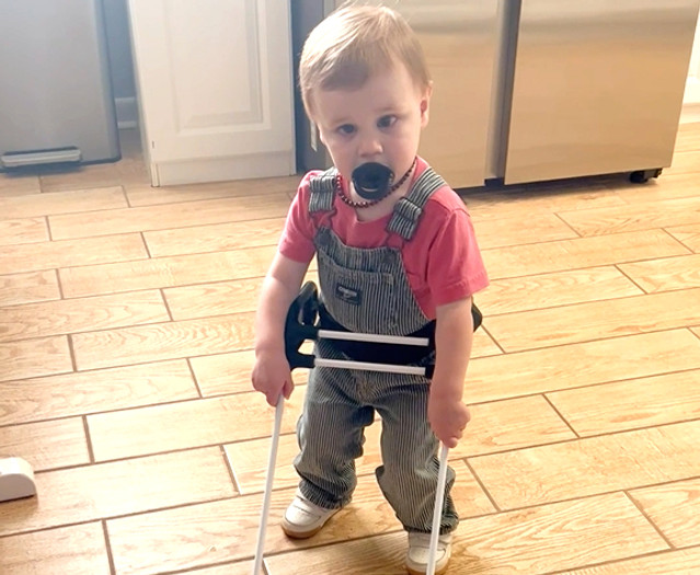 a blonde 1-year-old boy wearing a belt cane is standing in his kitchen wearing overalls and a red shirt with a pacifier in his mouth.