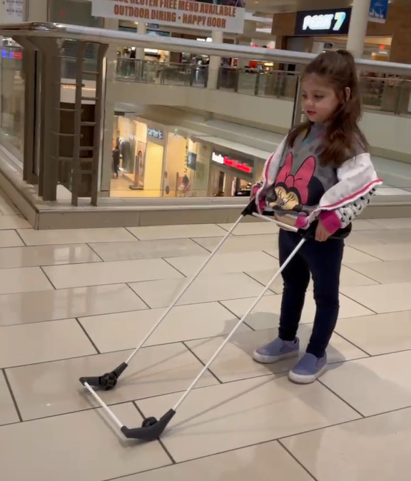 Young girl in a mall wearing a Belt Cane on tiled floor, wearing a Minnie Mouse shirt; railings and a Post 7 sign behind her.