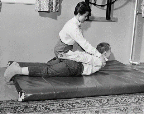 black and white photo of a child lying on his belly on a thick mat. His hands are clasped behind him, the PT kneels beside him and is pulling his shoulders away from the floor.