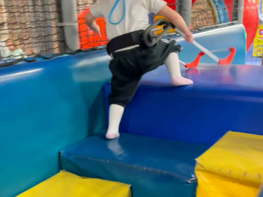 Child wearing a Belt Cane climbs colorful padded steps in a play area, wearing a white shirt and black pants, with a playful setting.