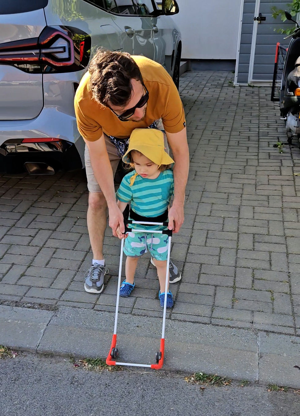 Parent crouching beside a child with a Belt Cane, both smiling near the drop off at the edge of the driveway.