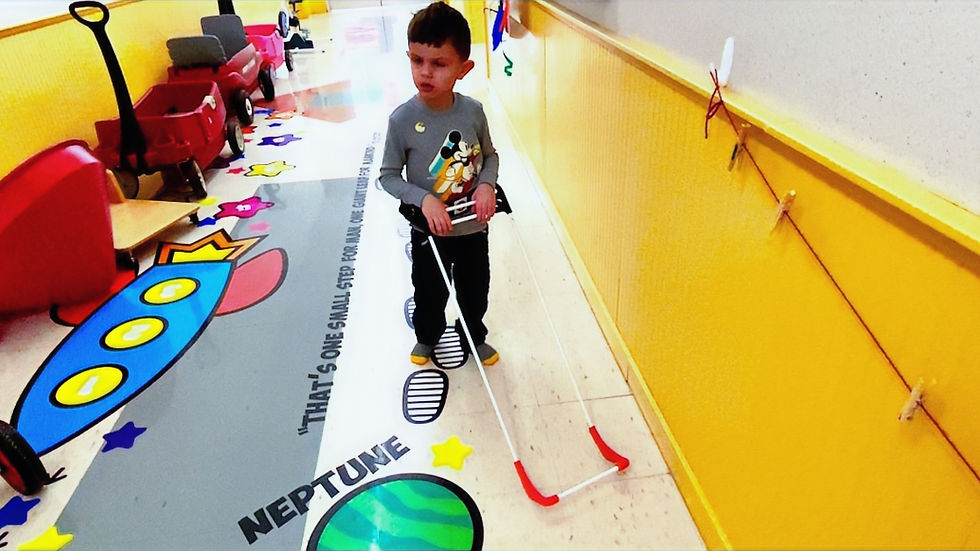 Young boy in a colorful hallway wearing a red and white belt cane. Floor has planets and footprints with text: "That's one small step..."
